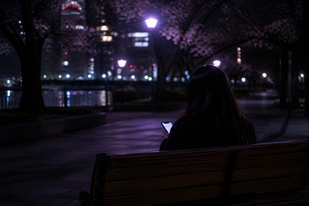 夜の公園のベンチでスマートフォンを見る女性の後ろ姿