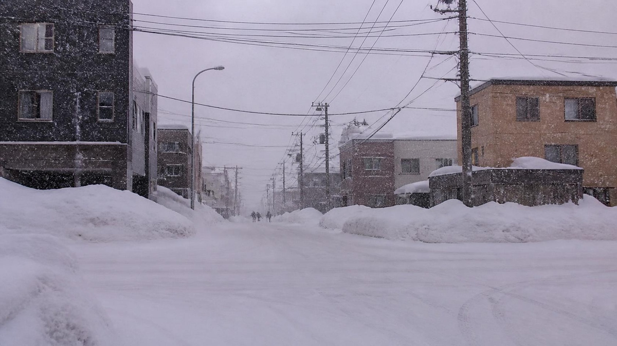雪が降りしきる昼の街角と、雪にかすむ遠くの人影